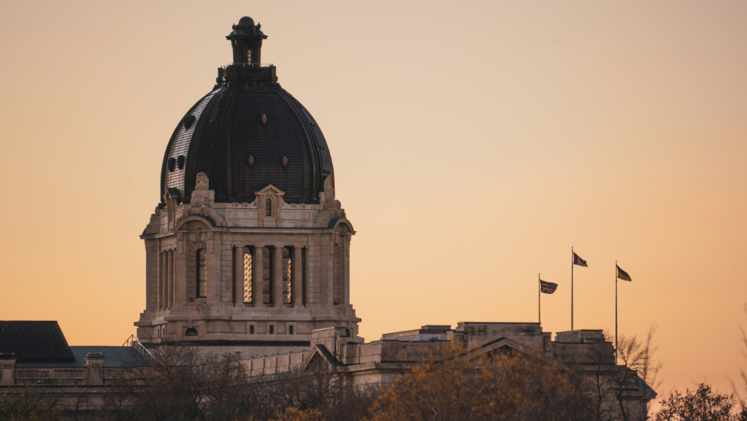 A domed stone government building is shown at sunset. Three flags are flying on flagpoles to the right of the dome. Trees with autumn foliage partially obscure the lower part of the building.