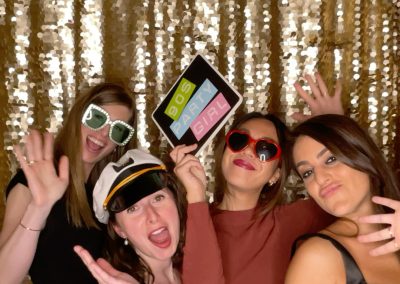 Four women pose in front of a gold sequin backdrop. One holds a sign reading “90s PARTY GIRL.” They wear outfits including heart-shaped sunglasses and a captain's hat, all smiling and waving at the camera.