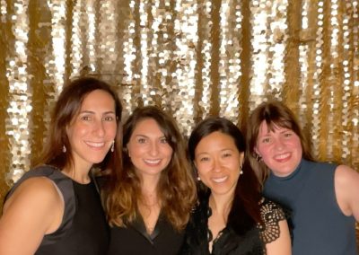 Four women stand together smiling in front of a gold sequin backdrop. They are dressed in semi-formal black and dark-colored outfits and are posing closely for the photo.