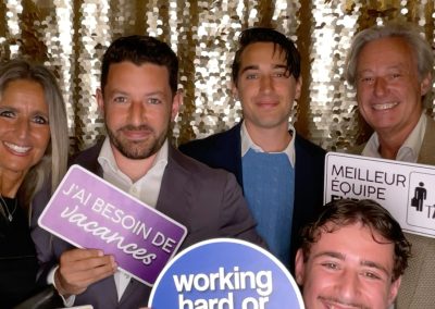 Five people pose in front of a gold sequin backdrop, holding various signs with messages like “working hard or hardly working?” and “J'ai besoin de vacances.” All are smiling and looking at the camera.