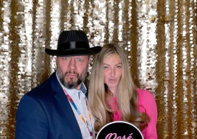 A man in a dark suit and hat stands next to a woman in a pink outfit holding a sign that says "Rosé LIFE." They pose in front of a gold sequin backdrop.