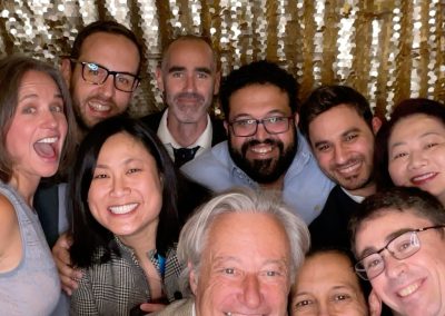 Ten adults smiling and posing closely together in front of a gold sequin backdrop. The group appears to be at a social event or celebration.
