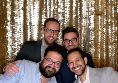 Four men pose together in front of a gold sequin backdrop. Three are wearing suits and glasses, while the fourth, in a light blue shirt, stands in front making a peace sign with his hand. All are smiling at the camera.