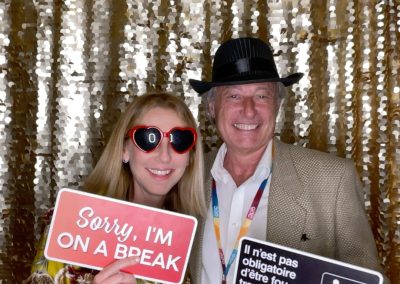 Two people stand in front of a gold sequin backdrop. The woman wears heart-shaped sunglasses and holds a sign reading "Sorry, I'm ON A BREAK." The man in a hat holds a sign with French text about taking breaks at work.
