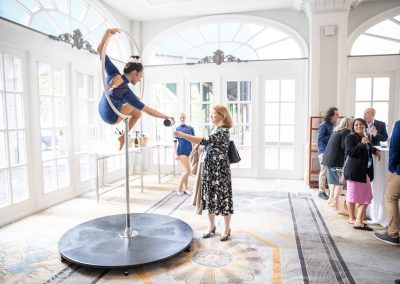 A woman in a patterned dress hands a drink to a pole dancer performing a split on a pole, inside a bright room with large windows. Several people are gathered around a table in the background.