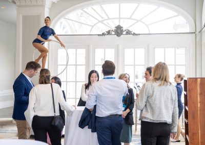 A group of people stand and talk around a cocktail table at an indoor event while a performer balances on a suspended hoop in the background near large windows.