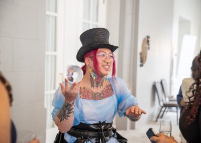 A person with pink hair, glasses, and visible tattoos wears a black top hat and a light blue dress, holding a clear sphere while smiling and talking to others at an indoor event.