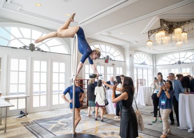 A performer in a blue leotard balances upside down on a vertical pole in a bright, elegant room while attendees watch and take photos during an indoor event.