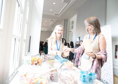 Two women wearing blue lanyards smile as they select candy from clear containers at a brightly lit indoor event with large windows and other attendees visible in the background.