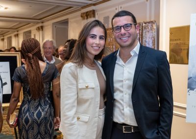 A woman and a man stand side by side, smiling at the camera at an indoor event. Several other people are visible in the background, along with signage and decorative elements on the walls.