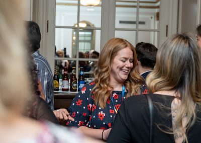 A woman with reddish-brown hair in a floral dress smiles while interacting with others at a social gathering in front of a bar with assorted bottles. Several people stand around her in a well-lit room with a mirror.