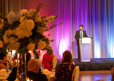 A man stands at a podium speaking on stage in a banquet hall with colorful lighting. People sit at round tables, some with floral centerpieces, listening to the speaker.