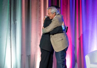 Two men in suits hug each other on a stage with multicolored vertical curtains and a white chair visible on the right side of the image.