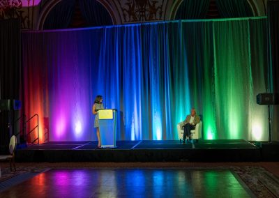 A woman stands at a podium on stage speaking, while an older man sits in a chair to the right. The stage has a multicolored, illuminated curtain backdrop, and the scene is set in a large, ornate room.