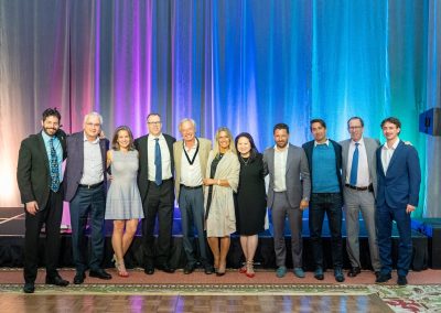 A group of twelve people, dressed in formal and semi-formal attire, stand side by side and pose for a photo in front of a stage with colorful blue, purple, and pink lighting.
