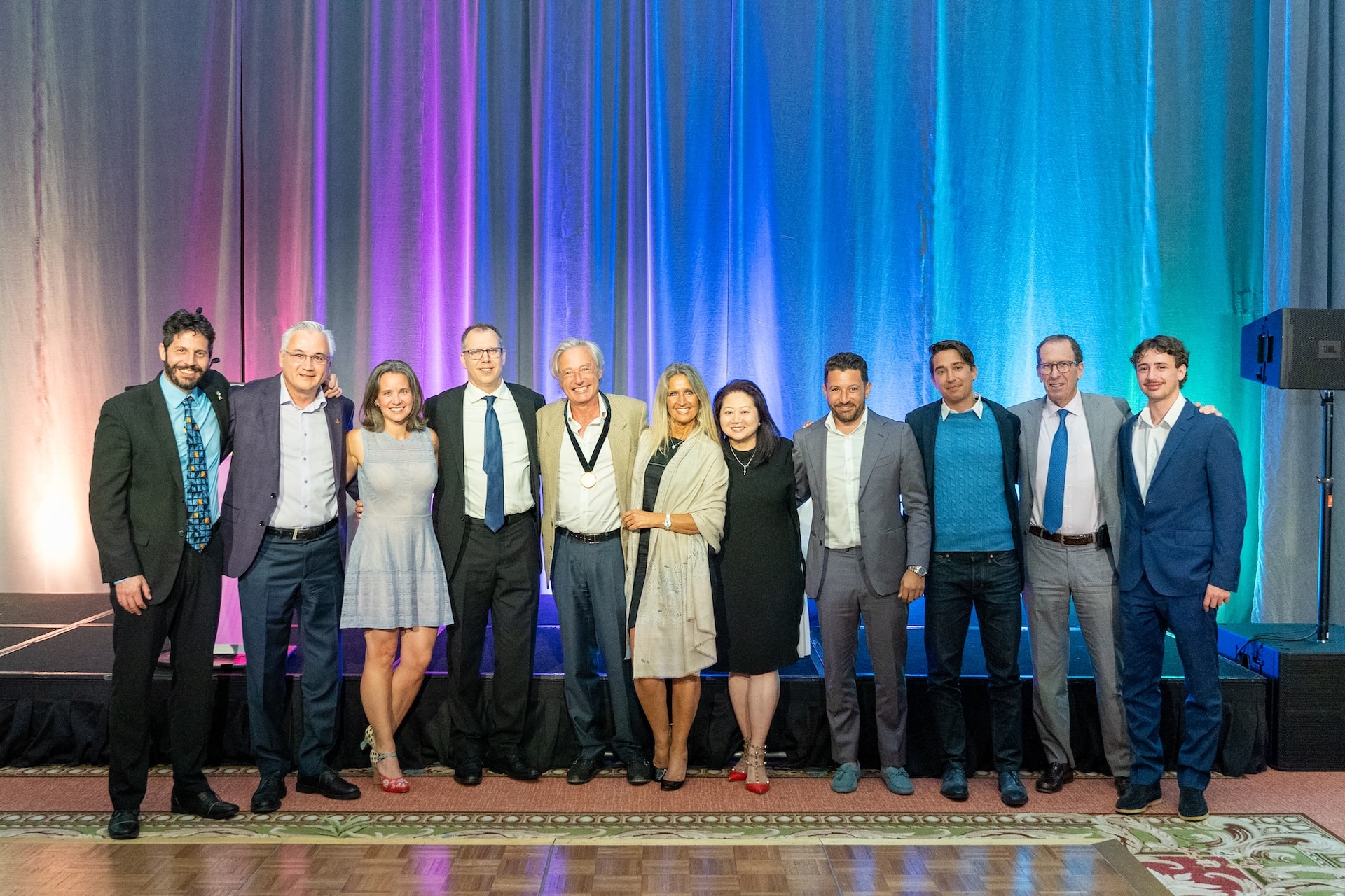 A group of twelve people, dressed in formal and semi-formal attire, stand side by side and pose for a photo in front of a stage with colorful blue, purple, and pink lighting.