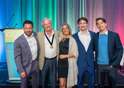 Five people stand together smiling in front of a colorful curtain and a podium with a “GOOD” sign. The man second from left wears a medal. All are dressed in business or semi-formal attire.