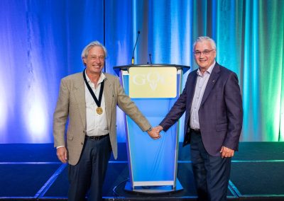 Two older men in suits stand in front of a podium with a "GQC" sign, smiling and holding hands. One man wears a medal around his neck. The background features blue and green curtains.