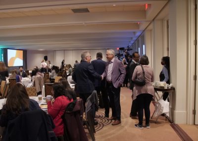 A group of people, some standing and some seated at round tables, attend a conference or meeting in a large room with a projector screen and refreshments on a side table.