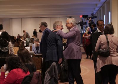 A group of people attend a conference or meeting in a large, well-lit room. Two men in suits greet each other with a handshake and a hug, while others sit at tables or stand and talk.