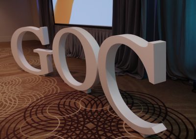Large white 3D letters spelling "GOC" are displayed on a patterned carpet in front of a gray curtain and a projection screen.