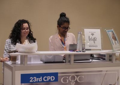 Two women stand behind a registration desk with laptops at an event. A sign on the counter displays wifi details. The desk has signage reading "23rd CPD" and "GOC." A coffee cup and brochures are also visible.