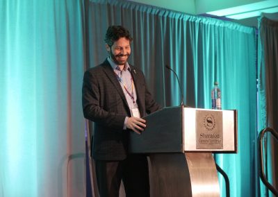 A man in a suit stands at a podium with a microphone and a water bottle, smiling, in front of teal curtains at a Sheraton Centre Toronto event space.