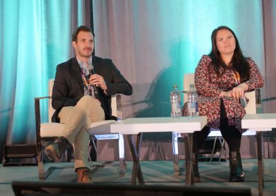 A man in a suit and a woman in a patterned dress sit on stage in armchairs with microphones. Two water bottles and glasses are on the table between them. Turquoise lighting covers the curtain behind them.