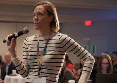 A woman with a striped shirt stands holding a microphone, speaking at an indoor event or conference. People are seated and working in the background.
