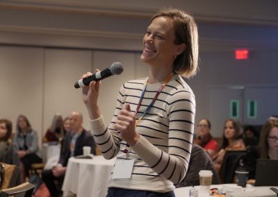 A woman in a striped shirt stands holding a microphone, smiling and speaking at an indoor event. Other attendees are seated at round tables in the background, listening.