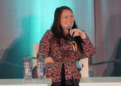 A woman with long brown hair and glasses, wearing a floral dress, speaks into a microphone while seated on a stage. Two water bottles are on the table in front of her. A teal and gray backdrop is behind her.