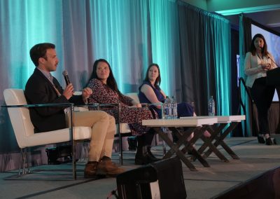 Four people are participating in a panel discussion on stage. Three are seated in chairs with microphones and water bottles, while one stands at a podium speaking. The background features teal curtains.