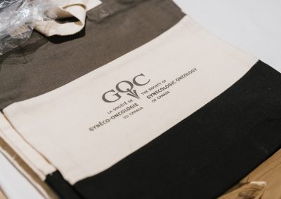 A beige and black tote bag with “The Society of Gynecologic Oncology of Canada” logo and text printed in English and French is placed on a table. A partially visible plastic item is near the top left.