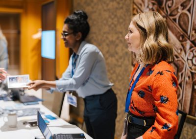 Two women stand behind a registration table with laptops and documents. One woman wears a floral red blouse and looks to the side, while the other, in a blue shirt, hands a paper to someone off-camera.
