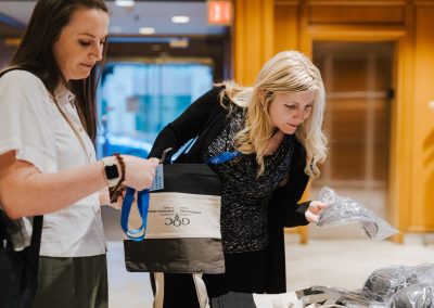 Two women stand at a table indoors, examining tote bags and items in plastic bags. One holds a lanyard and a tote bag, while the other looks at something on the table. The setting appears to be a conference or event.