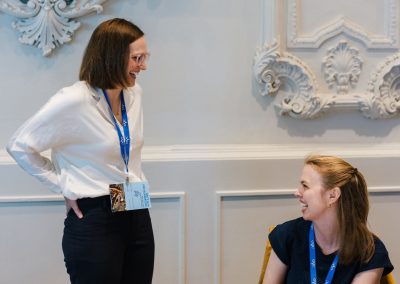 Two women wearing name badges and blue lanyards are talking and smiling in a room with ornate white wall decorations; one is standing and the other is seated.