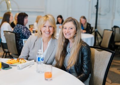 Two women sit next to each other at a round table set with drinks and plates of food during an indoor event. Other people are seated at tables in the background. Both women are smiling at the camera.