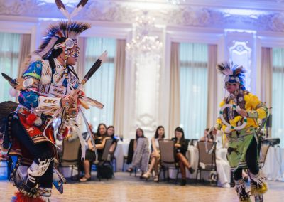 Two dancers in colorful, traditional Native American regalia perform indoors on a parquet floor, while several seated people in modern clothing watch in the background. The room is ornate with large windows and chandeliers.