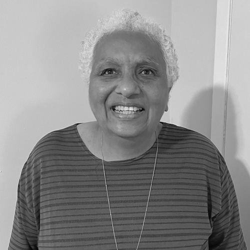 An older woman with short curly hair is smiling at the camera. She is wearing a striped top and a necklace, standing in front of a plain wall. The photo is in black and white.