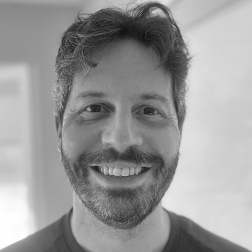 A man with wavy hair and a beard smiles at the camera. The background is indoors and softly blurred. The image is in black and white.