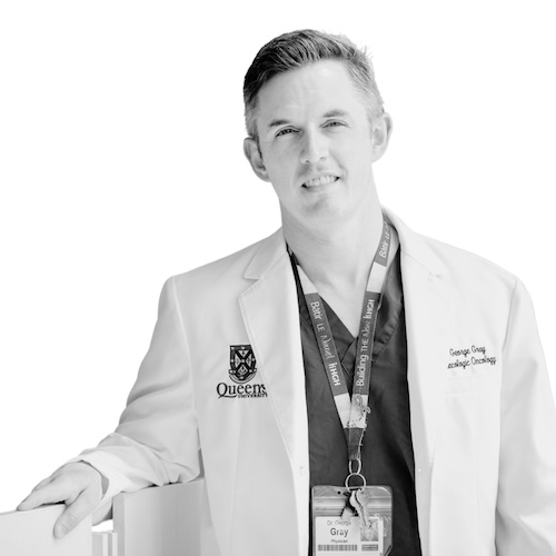 A man wearing a white lab coat with hospital badges stands in front of a white background, resting one hand on a railing. The lab coat features an embroidered logo and text. The image is in black and white.