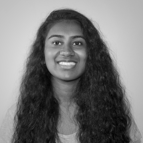A young woman with long, curly dark hair is smiling at the camera. She is wearing a light-colored top and is posed in front of a plain, light background. The image is in black and white.