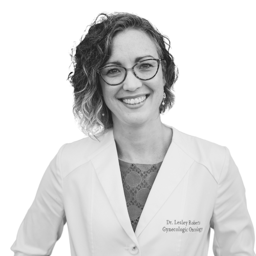 A woman with curly shoulder-length hair and glasses smiles at the camera. She is wearing a white medical coat with the name "Dr. Lesley Roberts, Gynecologic Oncology" embroidered on it.