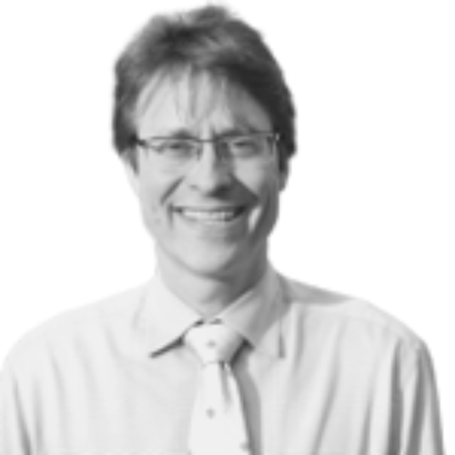 A man with short, dark hair and glasses is smiling. He is wearing a light-colored shirt and a tie. The background is plain white.