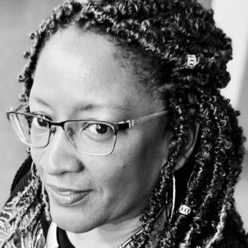 Black and white close-up photo of a woman with glasses, braided hair, and patterned accessories, looking slightly to her left with a neutral expression.