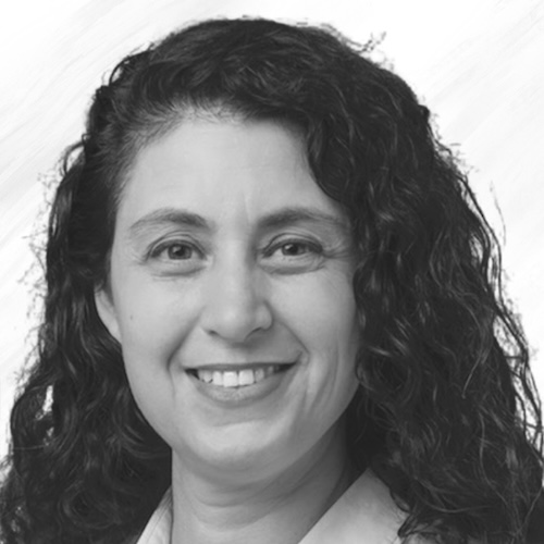 A woman with long, curly dark hair smiles at the camera. She is wearing a light-colored collared shirt. The background is plain and light in color. The image is in black and white.