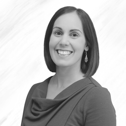 A woman with straight, shoulder-length dark hair smiles at the camera. She is wearing a textured, long-sleeve top and drop earrings. The background is plain and light-colored. The image is in black and white.
