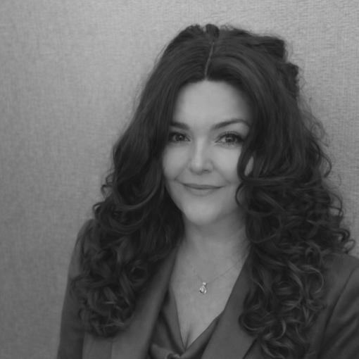 A woman with long, dark, curly hair is smiling slightly while facing the camera. She is wearing a blazer and a necklace. The background is plain and textured. The image is in black and white.