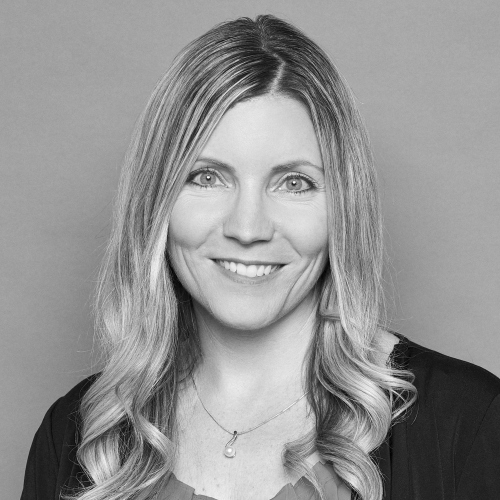 A woman with long, wavy, light-colored hair is smiling at the camera. She is wearing a dark top and a necklace. The background is plain and light-colored. The image is in black and white.